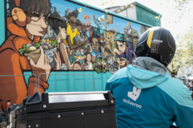 View from behind a Deliveroo motorcycle delivery person looking at a colourful mural of Bristol fast food outlets painted onto a large wall space in the city.