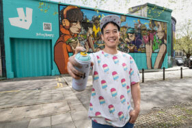 Graffiti artist Silent Hobo is looking to camera and has his right arm raised to the lens, in his hand is a spray can and his finished mural is behind him on a building wall.