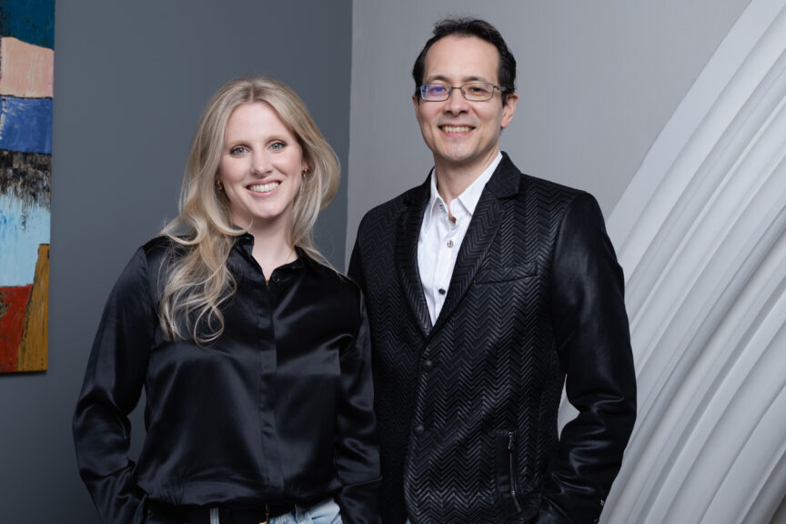 Blonde female and brunette male wearing glasses, both dressed in smart black tops, smiling to camera for a business portrait.