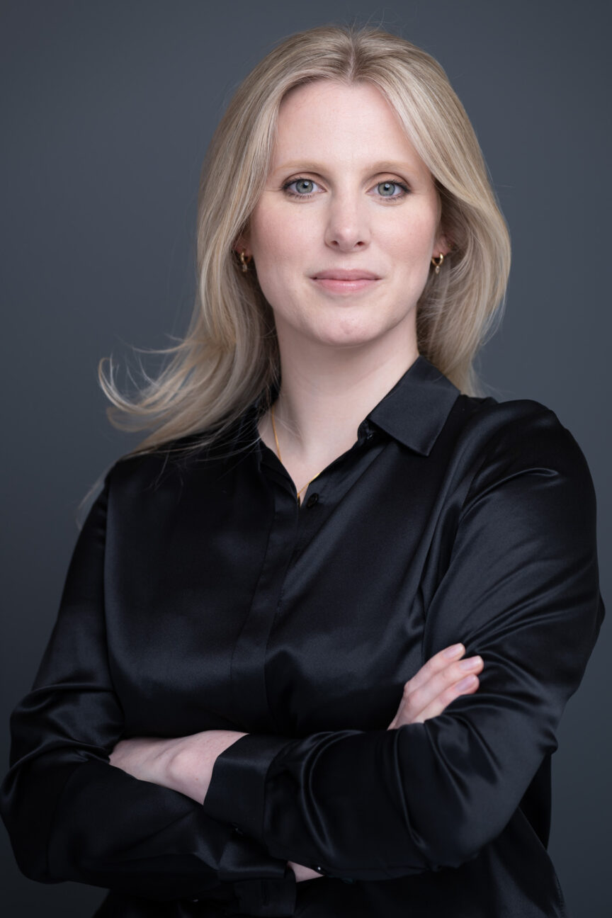 Blonde female dressed in a smart satin black blouse, smiling to camera for a business portrait.