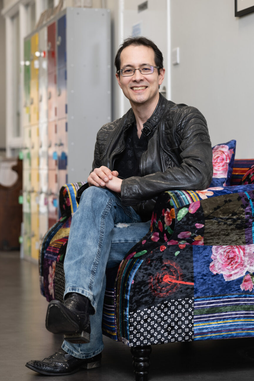 Paul Albone is seated on a colourful sofa. He's smiling to camera, wearing a black leather jacket, blue jeans and stylish black boots.