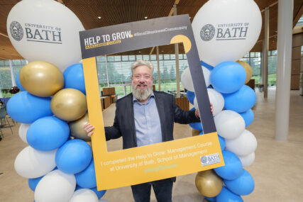 A grey-haired, bearded man in a suit stands smiling while holding a large cardboard Instagram-style frame. He is flanked by two towers of balloons.