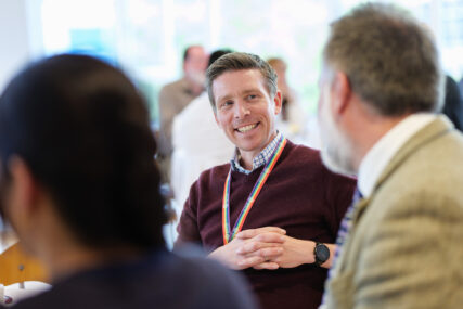 A man wearing a multi-coloured PRIDE lanyard is smiling to the person seated next to him.