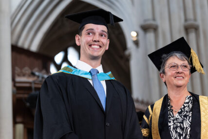 A University of Bath graduate stands smiling broadly at the audience moments after receiving his graduation. He is standing next to the Pro-Chancellor and the background is the interior of Bath Abbey.