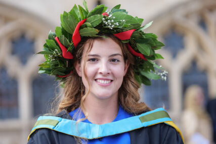 A female University of Bath graduate looks directly to camera and smiles. She is wearing a laurel wreath on her head, a tradition of Italian students.