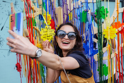 A young woman in sunglasses smiles to camera while holding her phone at arm's length for a selfie in front of colourful ribbons. She's holding a message of support she wrote on a plastic yellow flower tile.