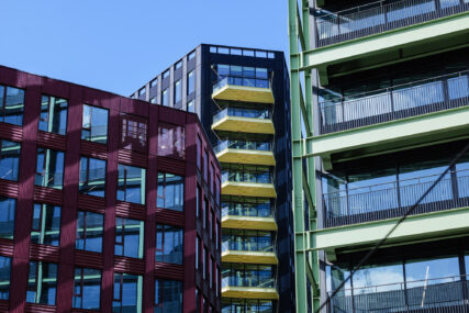 Sections of three modern office buildings shown together in a 'compressed' view using a long lens. From left, a burgundy-coloured building, the centre building has yellow accented balconies and the righ-hand building features green external steel girder construction.