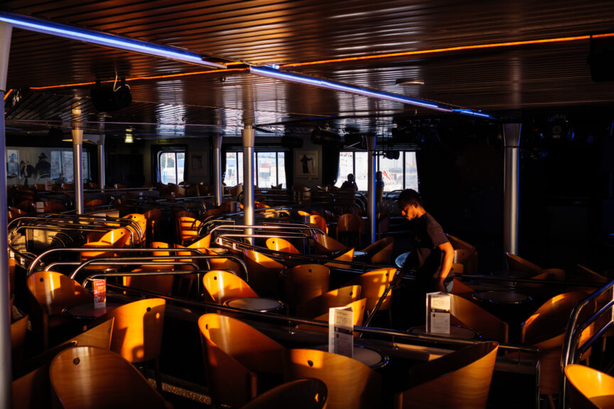 Golden evening sunlight illuminates the curved wooden chairs on board the Portsmouth to Saint Malo ferry as a cleaner moves through, vacuuming the floor.