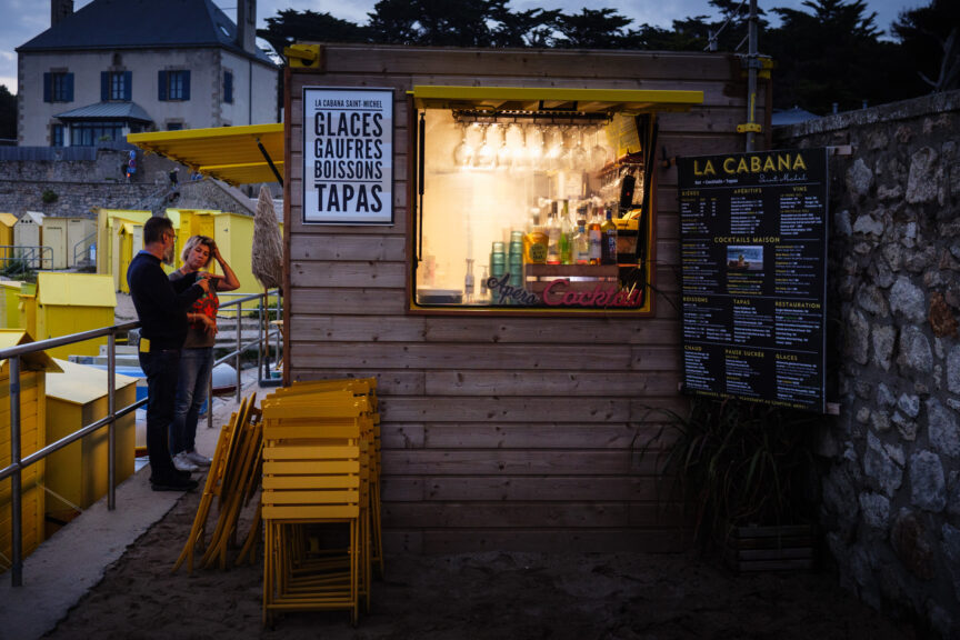 Small snack cabin and bar in evening light at Batz-sur-Mer, Brittany, as a man and a woman stand outside chatting.