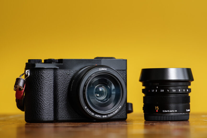 A panasonic Lumix GX9 camera on a wooden table with a yellow background. The camera has the Panasonic 20mm lens fitted and is shown next to a Lumix/Leica 15mm lens.