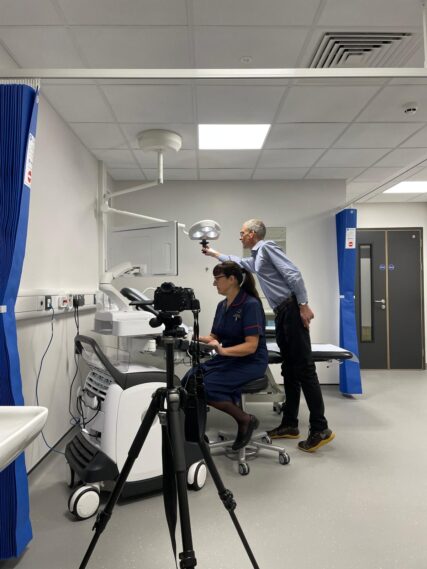 A medical nurse sits at a electrocardiogram (ECG) machine while a photographer adjusts the medical light behind her. The photographer's camera is on a tripod in the foreground.