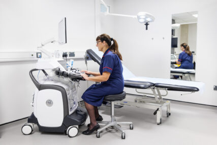 A clinical nurse sits at an electrocardiogram (ECG) machine, looking at the screen and operating the controls. There is an examination bed in the background.