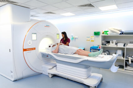 A male patient is on the MRI bed wearing a hospital gown and the head scanner piece. A female nurse is standing the other side of the bed operating the controls.