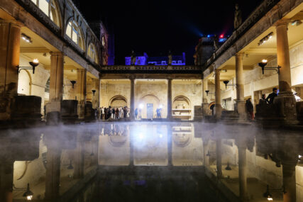 It's night time and steam rises from the naturally heated waters of the Roman Baths in Bath, England. The surrounding stone structures are illuminated as people can be seen milling about.