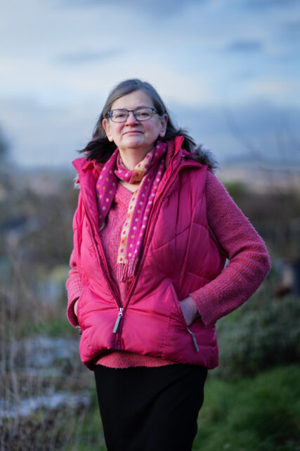 Outdoor portrait of Katharine Tylko in a nature setting. She is wearing a pink coat, scarf and blouse and looking to camera.