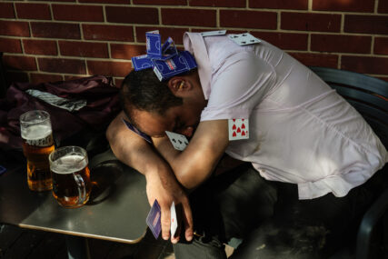 A man sits slumped, sleeping with his head on his arms at an outdoor bar table with half-drunk glasses of beer near him. For a joke, his drinking buddies have covered him in playing cards, inserted them between his fingers, stacked them on his head and put a cigarette butt in his ear.