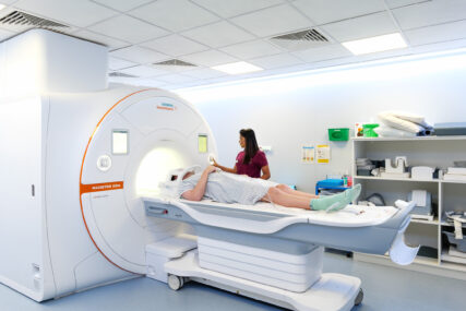 A patient is lying on a MRI machine bed, about to be slid in for a scan, while a medical nurse operates the controls.