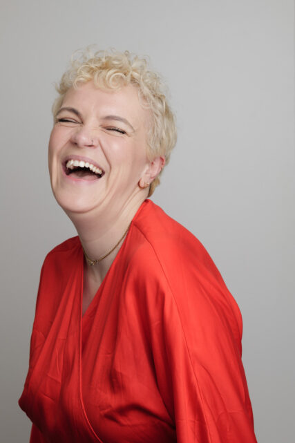 A woman with short blonde hair wearing a bright crimson blouse laughs.