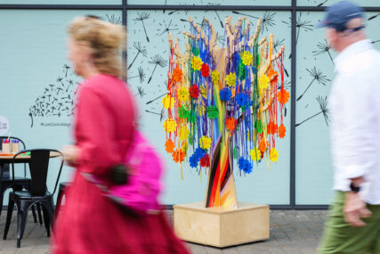 A small tree made of polished plywood is covered in colourful ribbons and discs which have messages of support for the LGBTQ+ community written on them. Two shoppers are blurred walking past the tree.