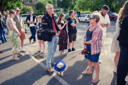 Coldplay fans queue in Frome's Cheese & Grain car park. One woman has a small dog on a lead. The dog is wearing a blue cone round its neck.