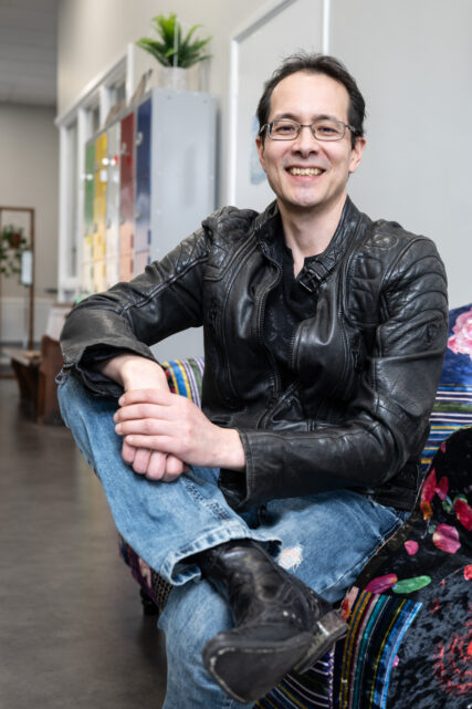 Portrait of a caucasian man in black leather jacket, black leather boots, glasses and jeans sitting casually on a sofa, smiling to camera.