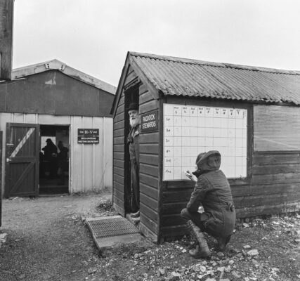 A bearded race steward stands in the doorway of the Stewards; hut at Larkhill point-to-point while a female official crouches to write up the latest scores on w whiteboard which is affixed to the outside wall of the hut. He is wearing bowler hat and tweeds, she is in wellies and raincoat. The weigh-in room building is visible beyond.