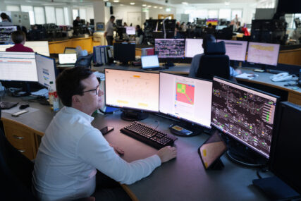 A man in smart office shirt and glasses looks to a bank of three monitors showing rail movements and reports. An open-plan office of more workers and screens is visible beyond.