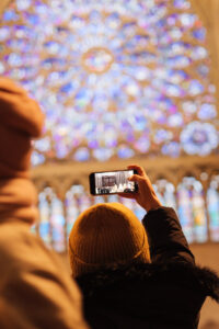 Rear view of a female tourist in an orange knitted hat raising her iPhone to take a photo of a holy mass underway in Notre Dame Cathedral, Paris, with one of the beautifully restored circular stained glass windows visible out of focus beyond.