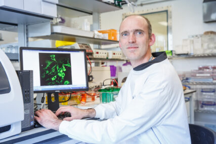 A male scientist in white lab coat is turning away from his computer monitor to look beyond the photographer. There is a micoscope image of a sample on his computer monitor.