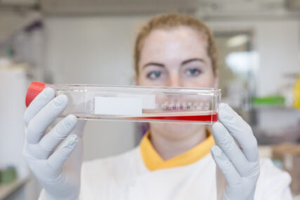 A female scientist looks at a red coloured liquid in a large test tube which she is holding horizontally between her face and the camera while wearing PVC gloves.