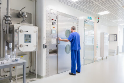 A scientist in blue scrubs stands looking into a large steel door with round porthole windows. He's in a mostly white corridor with other scientific equipment installed to his left.