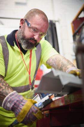 A bearded man in safety glasses and high-visibility vest and thick gloves looks down on a shaped sheet of steel he is holding and cutting.