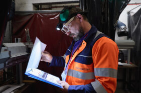 A bearded male bus engineer in goggles, hat and high-vis vest shines his head torch on a set of instructions so he can read them in a welding bay of a workshop.