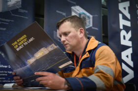 A mechanic in orange and blue overalls reads a pamphlet on Varta batteries, There is a large Varta sign just behind him.