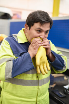 A mechanic in yellow and blue overalls holds a thick yellow rubber glove to his mouth to blow air into it to test for any leaks.