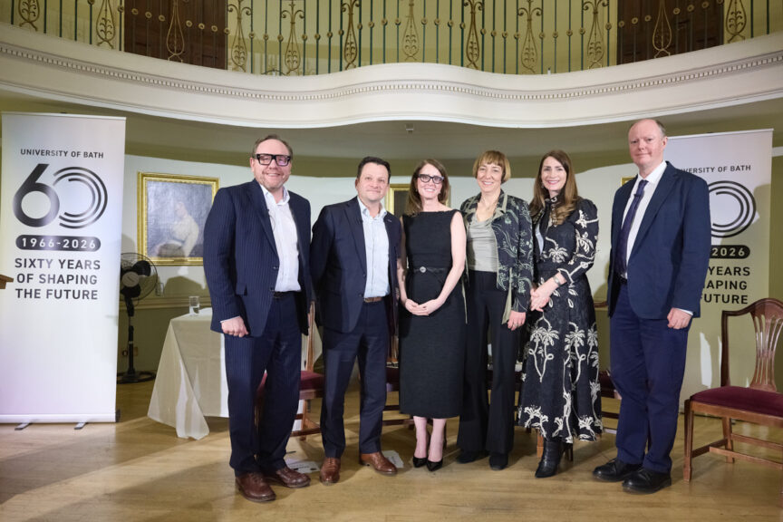 A group of six people (three men, three women) standing in a row, wearing formal evening attire in an ornate room are looking to camera for a group photo. There are pop-up banners announcing the 60th anniversary of the University of Bath.