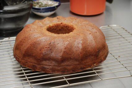 A circular cake with a hole through the middle sits on a wire cooling rack on a steel kitchen top.