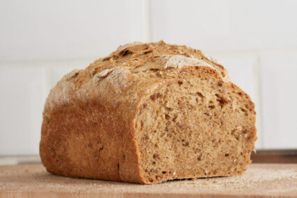 A cut loaf of bread on a wooden bread board with white tiles behind. The bread is dark as it's made with malted flour. We can see the texture and bubbles in the bread.