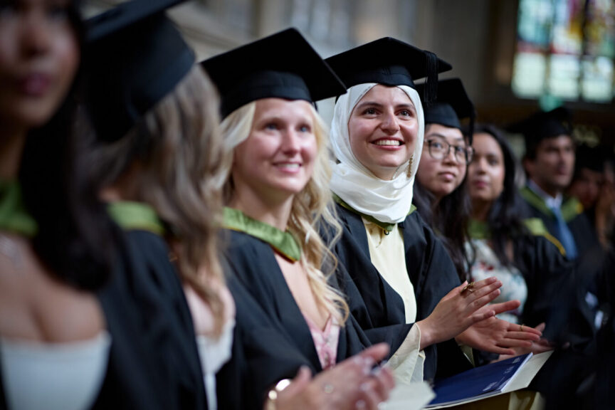 We are looking down a line of seated female students in the ornate surroundings of Bath Abbey. The students are in caps and gowns, looking up to a screen which which we can't see. The focus of the photo is on one student who in addition to her dark cap and gown is also wearing a head and neck scarf. She is smiling and clapping as fellow students take to the graduation stage (which we also can't see).